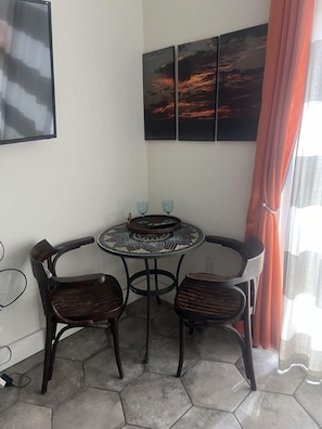 A corner room with a round table, two chairs, a wall-mounted TV, and a sunset painting.