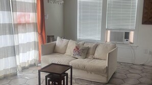 A living room with a sofa, a small table, hexagonal floor tiles, a window with blinds, and a wall-mounted air conditioning unit.
