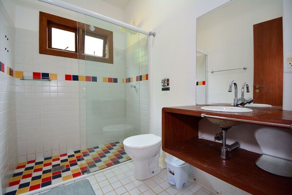 Un baño con cabina de ducha de vidrio, inodoro blanco, tocador de madera con lavamanos y piso de baldosas con un diseño colorido.