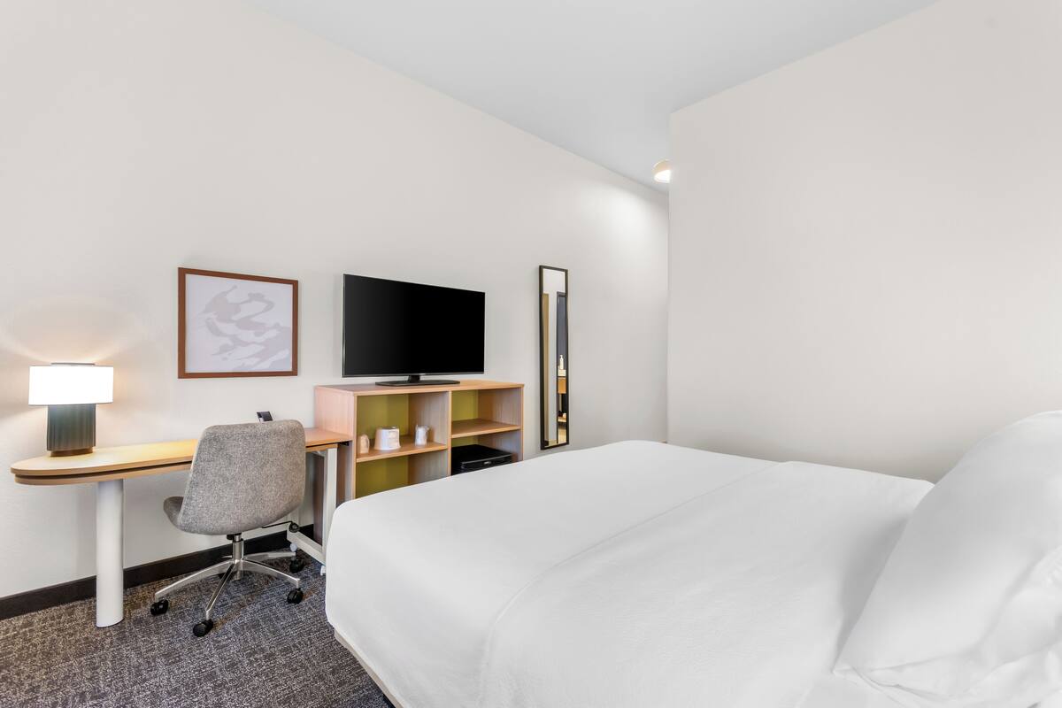A hotel room with a bed, a desk with a lamp, a chair, a television, and a shelf with items.