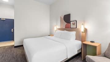 Une chambre d’hôtel avec un grand lit, une table de chevet avec une horloge, une chaise et une porte visible au fond.