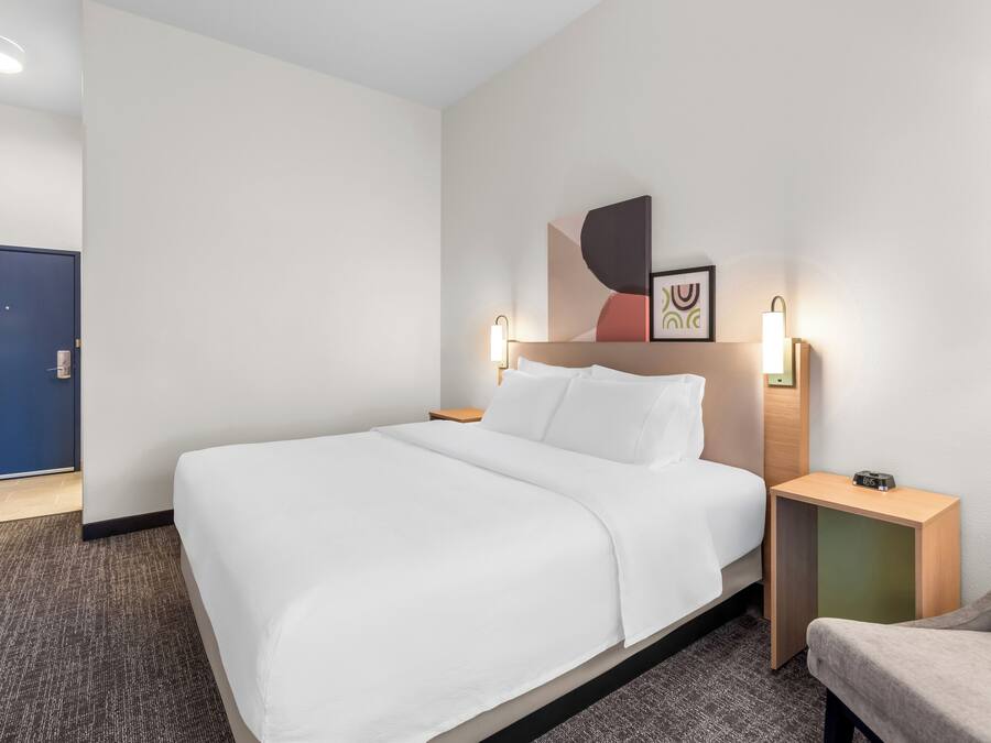Une chambre d’hôtel avec un grand lit, une table de chevet avec une horloge, une chaise et une porte visible au fond.