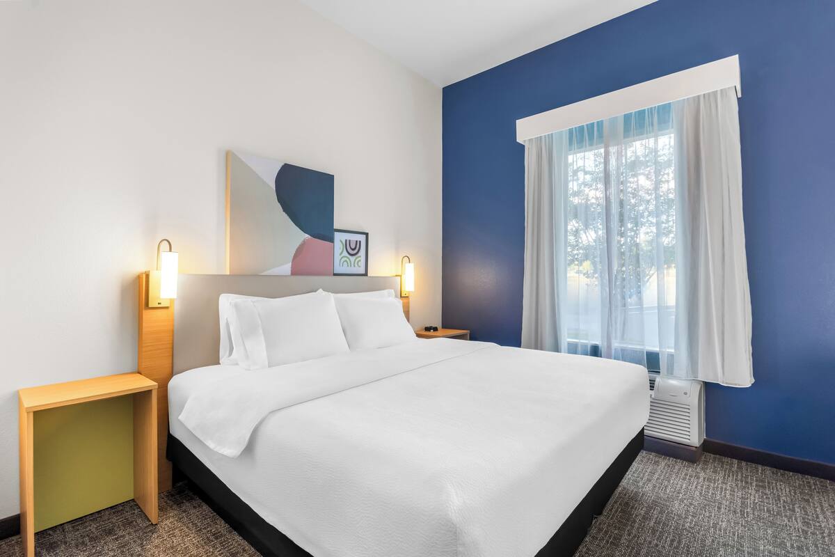 A neatly made bed with white linens, two bedside tables with lamps, a large abstract painting on the wall, and a window with sheer curtains.