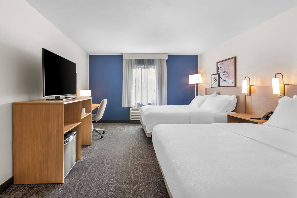 A hotel room with a TV, desk, two beds, a chair, and a window with curtains.
