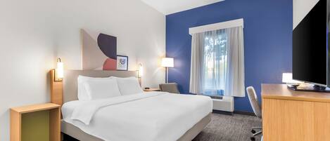 Una habitación de hotel con cama, mesitas de noche, escritorio, silla, televisor y ventana con cortinas.
