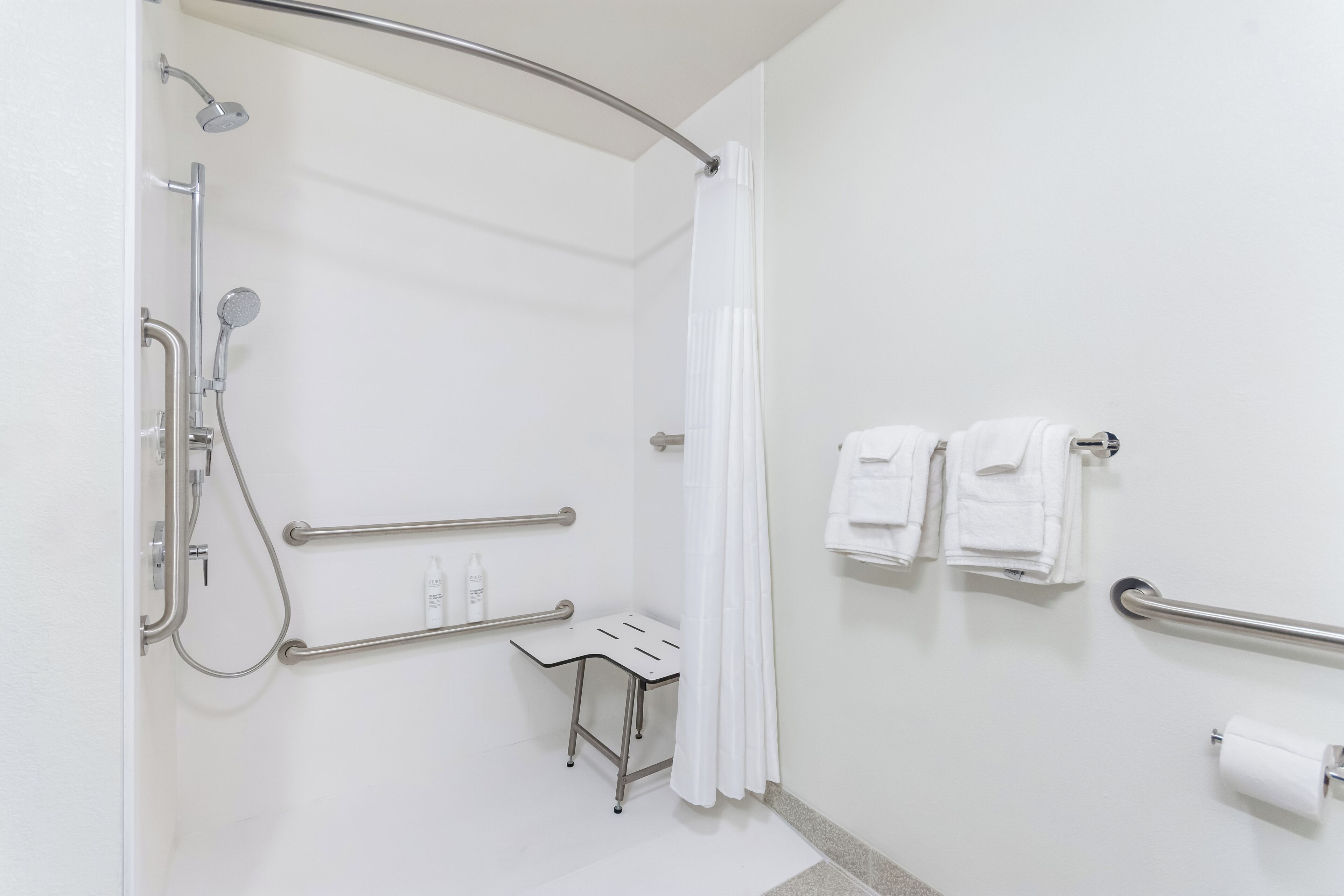 Ein modernes Badezimmer mit ebenerdiger Dusche, Haltegriffen, einem kleinen Tisch und einem Handtuchhalter.