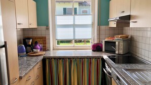 A kitchen with wooden cabinets, a window with blinds, a toaster, a microwave, and a colorful curtain. - Vacation apartment with terrace and garden (Piding)