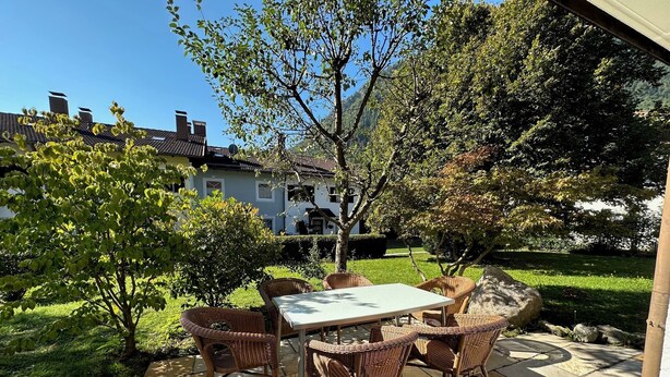 An outdoor patio with a table and wicker chairs, surrounded by greenery and a house in the background. - Vacation apartment with terrace and garden (Piding)