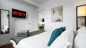 A modern hotel room with a large bed, a flat-screen TV mounted above it, a painting on the wall, and a small table with chairs in the background.