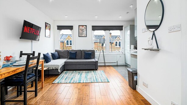 A modern living room with a wooden dining table, grey sofa, and a TV mounted on the wall.