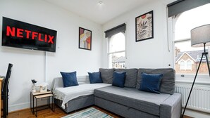 A modern living room with a grey sectional sofa, a flat-screen TV displaying 'NETFLIX', a small side table with a lamp, and artwork on the walls.