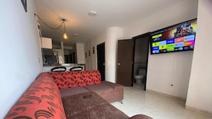 Una sala de estar con un sofá, un televisor montado en la pared y una zona de cocina al fondo.