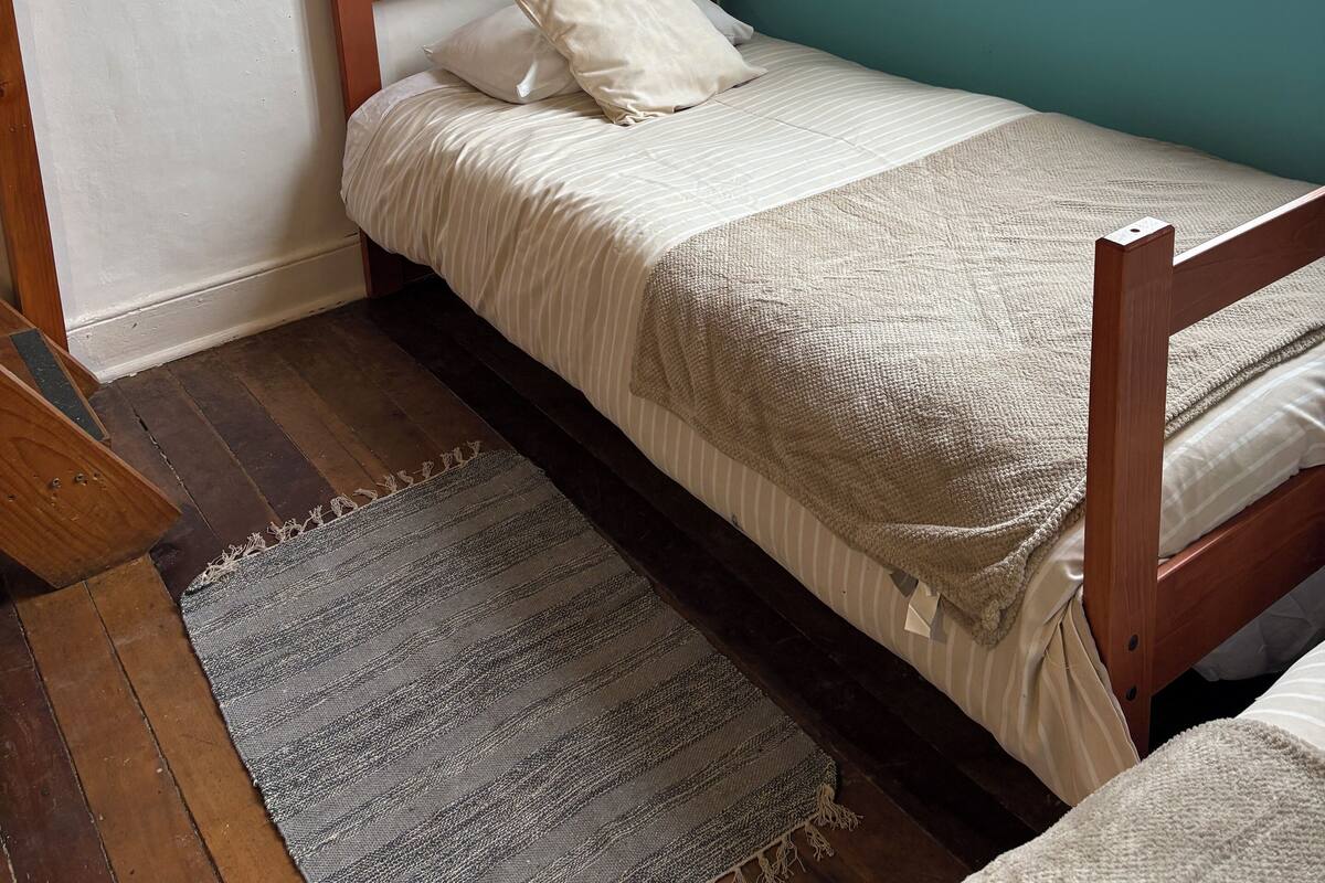 Ein Einzelbett mit Holzrahmen, einer gestreiften Bettdecke und einem beigen Kopfkissen. Ein grauer Streifen Teppich liegt am Fußende des Bettes.