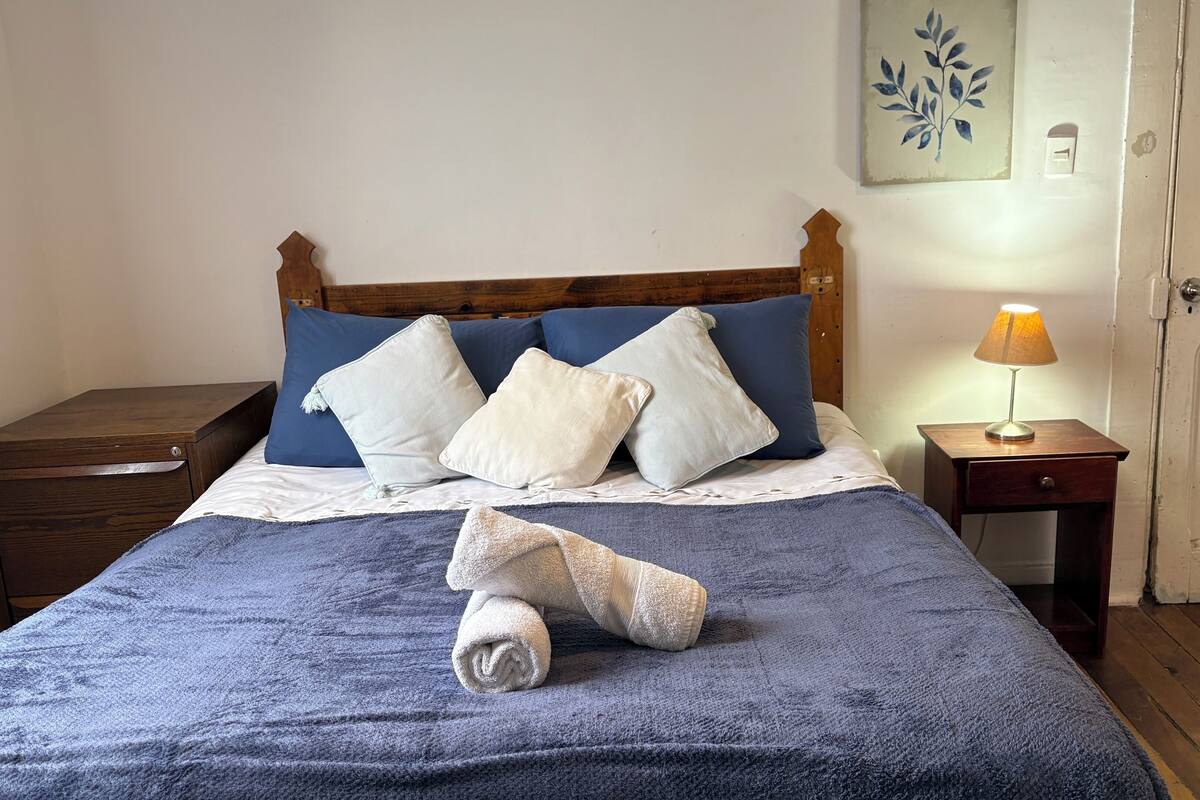 Ein ordentlich bezogenes Bett mit blauer Decke, weißen und blauen Kissen und einem aufgerollten Handtuch. Im Hintergrund ist ein Nachttisch mit Lampe, eine Holzkommode und eine Tür zu sehen.