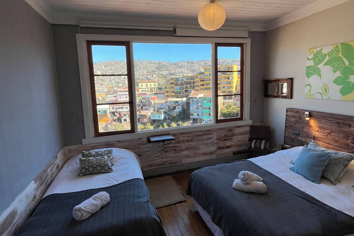 Ein Schlafzimmer mit zwei Betten, Holzboden, einem großen Fenster mit Blick auf die Stadt und einer Holzzieldecke.