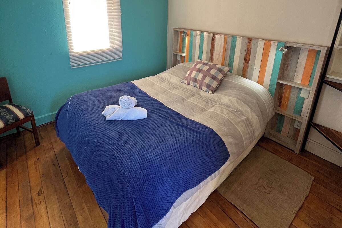 Ein Schlafzimmer mit Holzboden, einem Bett mit gestreifter Kopfteil und einem Fenster mit Jalousien sowie einem Sessel.