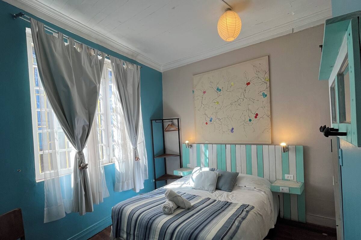 Ein Schlafzimmer mit Bett, einem Sessel, einem Nachttisch, einem Wandbild und einem Fenster mit Vorhängen.