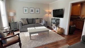 Un salón con un sofá gris, una mesa de centro blanca y suelo de madera. Hay un televisor montado en la pared y una lámpara sobre una mesita auxiliar.