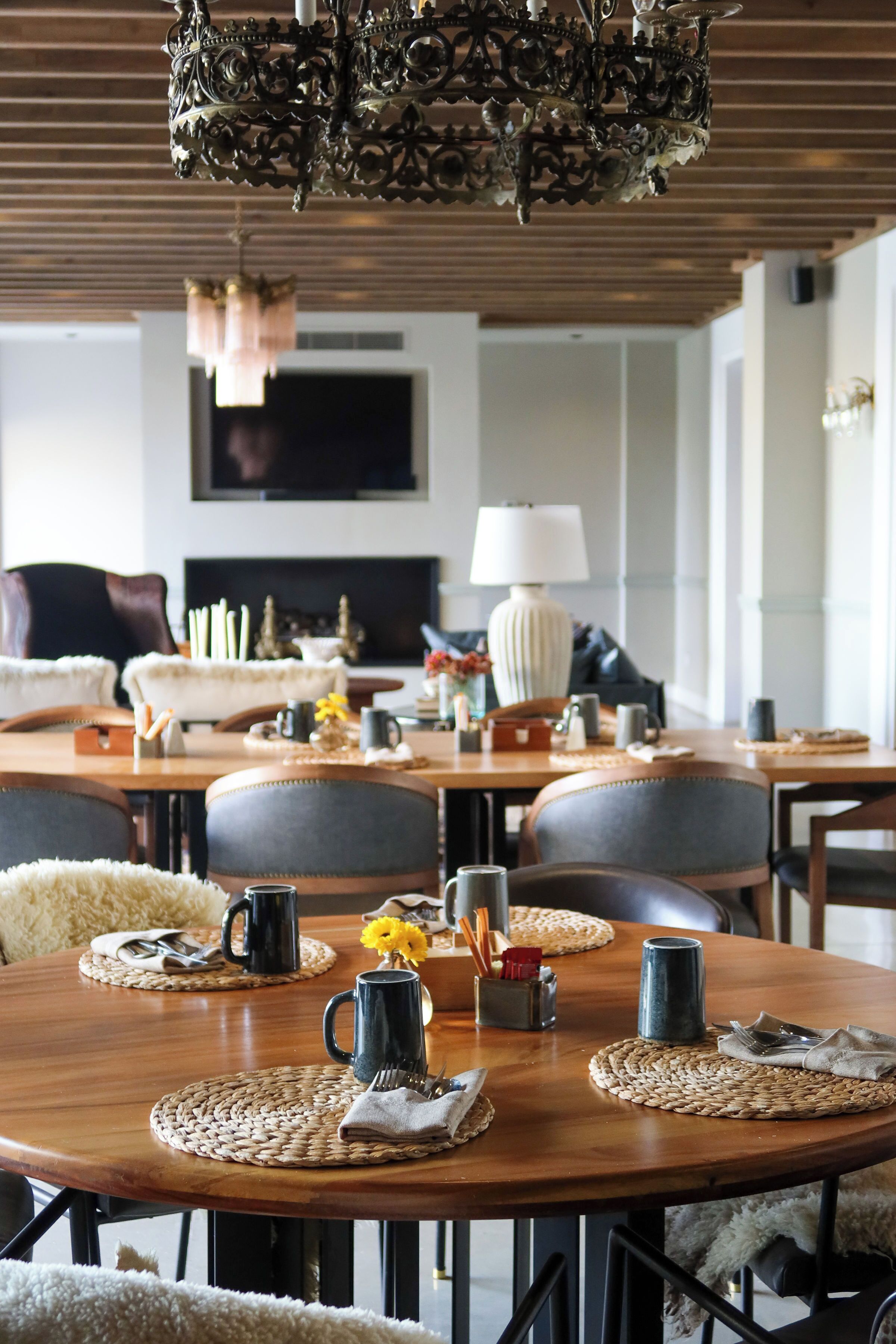 A dining area with a wooden table set for four, surrounded by upholstered chairs. A chandelier hangs above, and a television is mounted on the wall in the background.