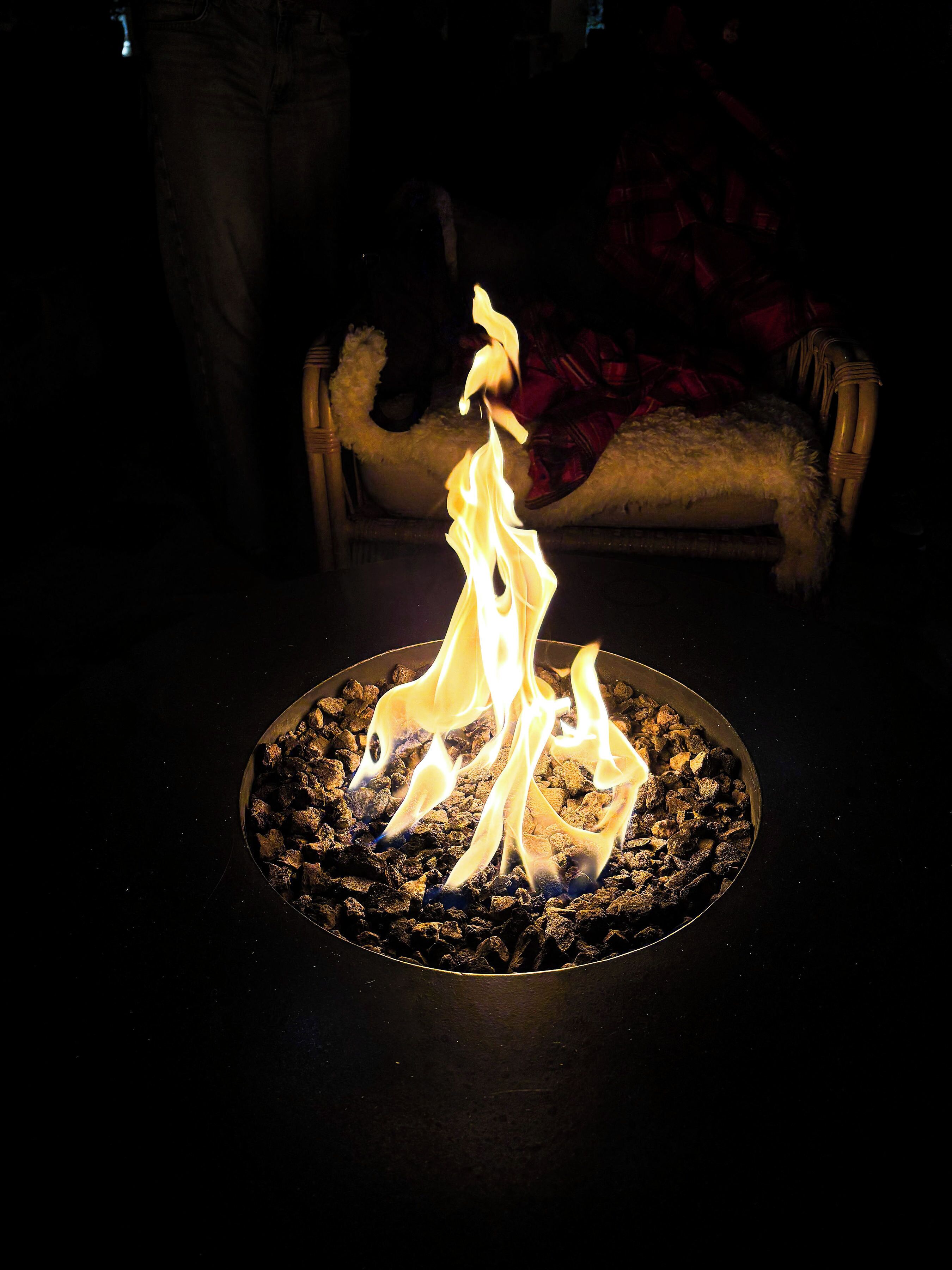 A fire pit with flames, surrounded by a dark room with a chair and a blanket in the background.