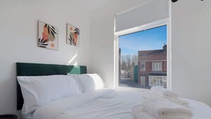 A bedroom with a bed, pillows, a green headboard, a window with a view of buildings, and two wall art pieces.