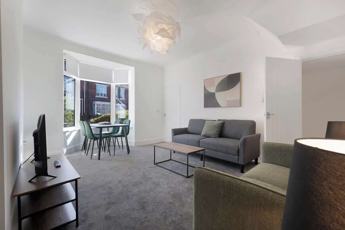A modern living room with a grey sofa, a coffee table, a TV on a desk, and a dining area with chairs and a table.