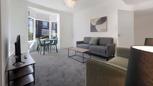 A modern living room with a grey sofa, a coffee table, a TV on a desk, and a dining area with chairs and a table.