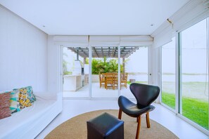 A modern living room with a sofa, a black chair, and a small ottoman. There is a glass door leading to an outdoor patio with a table and chairs. - Bz68 Centro Casa piscina priv 6 quartos (Buzios)