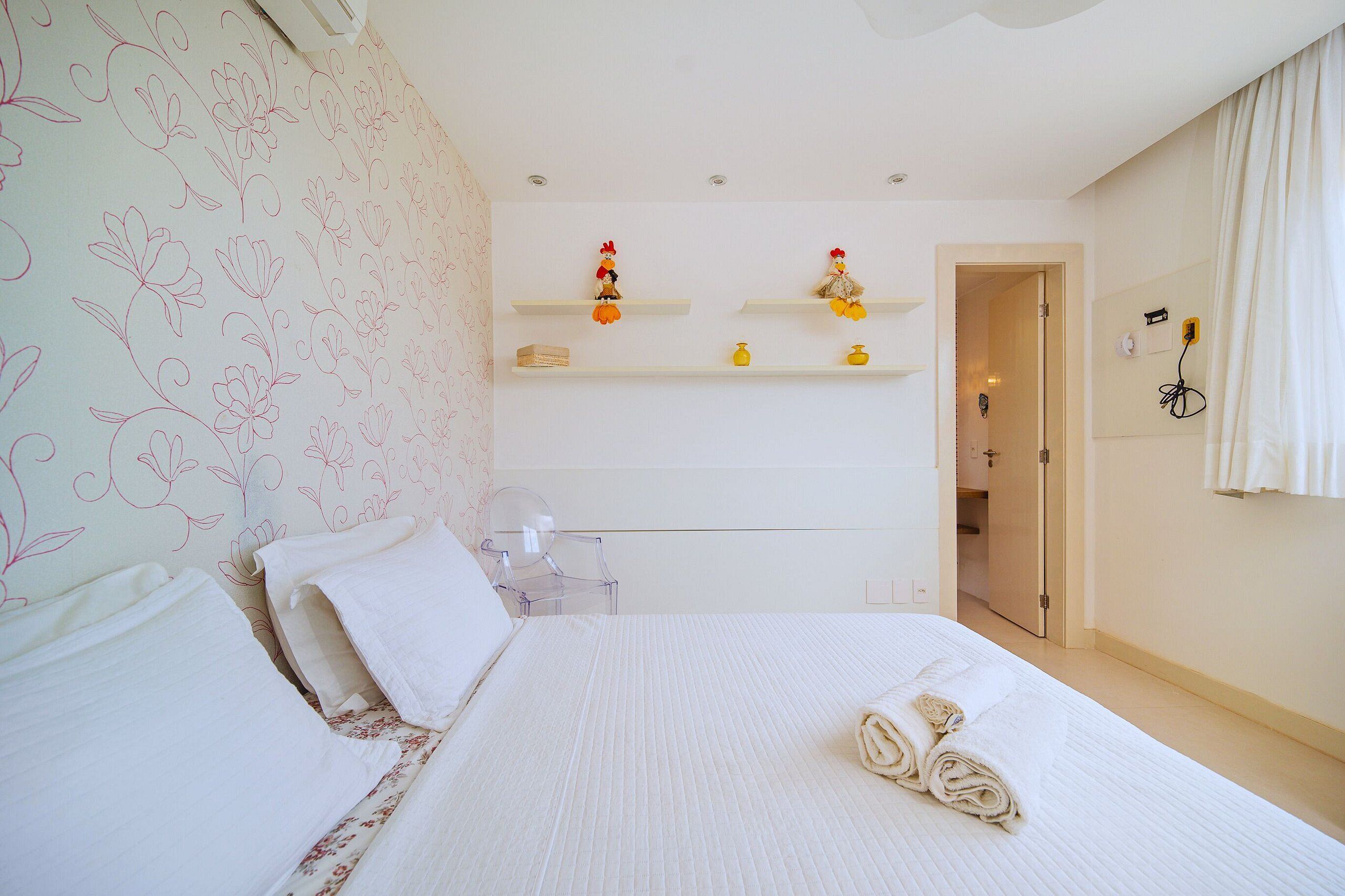 A neatly made bed with a rolled towel, a clear plastic chair, and two white shelves with decorative items.