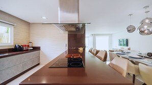 A modern kitchen with a central island, stainless steel appliances, and a dining area with a long table and chairs. - Bz68 Centro Casa piscina priv 6 quartos (Buzios)
