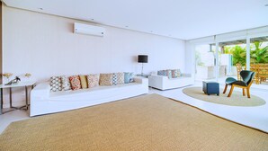 A modern living room with a white sofa, a coffee table, and a chair. There is a large window providing a view of the outdoors. - Bz68 Centro Casa piscina priv 6 quartos (Buzios)