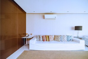 A modern living room with a white sofa, patterned cushions, a small side table, a lamp, and an air conditioning unit on the wall. - Bz68 Centro Casa piscina priv 6 quartos (Buzios)