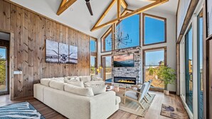A spacious living room with a large beige sectional sofa, a stone fireplace, and large windows offering a view of the outdoors.