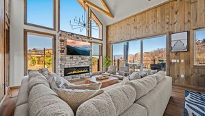 A spacious living room with a stone fireplace, large windows, and a wooden ceiling.