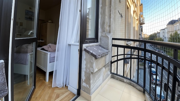 A balcony with a view of a city street, a window with a curtain, and a small seating area with cushions.