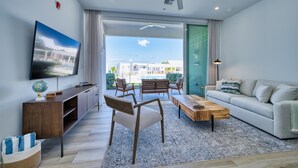 A modern living room with a flat-screen TV, wooden furniture, and a view of a pool and buildings. - Sunset Point: Chic Meets Luxury, Sophisticated Style Meets Ocean Serenity (West Bay)