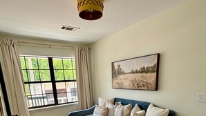 A room with a blue sofa, a small round table, a window with curtains, a ceiling light, and a painting on the wall. - Cedar Bucket Lofts - Cozy 2-bedroom apartment in Oxford! (Oxford)