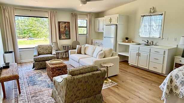 A spacious living room with a white sofa, two armchairs, a coffee table, and a dining area with a small table and chairs. There is a kitchen area with cabinets and a refrigerator. - Cedar Bucket Lofts - Cozy 2-bedroom apartment in Oxford! (Oxford)