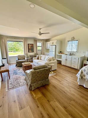 Ein geräumiges Wohnzimmer mit einem weißen Sofa, zwei Sesseln, einem Couchtisch und einem Essbereich mit einem kleinen Tisch und Stühlen. Es gibt eine Küchenzeile mit Schränken und einem Kühlschrank.