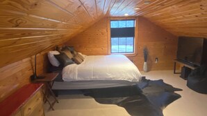 A cozy attic bedroom with a large bed, wooden walls, a window, a TV, and a dresser. - Cozy cabin with 1 bedroom, Lakefront views in tranquil Terry! (Terry)