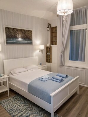 A bedroom with a bed, nightstands, a bookshelf, a chandelier, and a window with curtains. - Cracow Route #2 - Old Town (Kraków)
