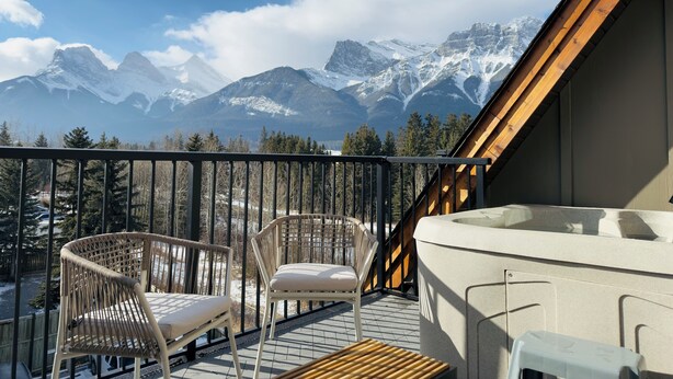 A balcony with two wicker chairs, a wooden table, and a view of snow-capped mountains.