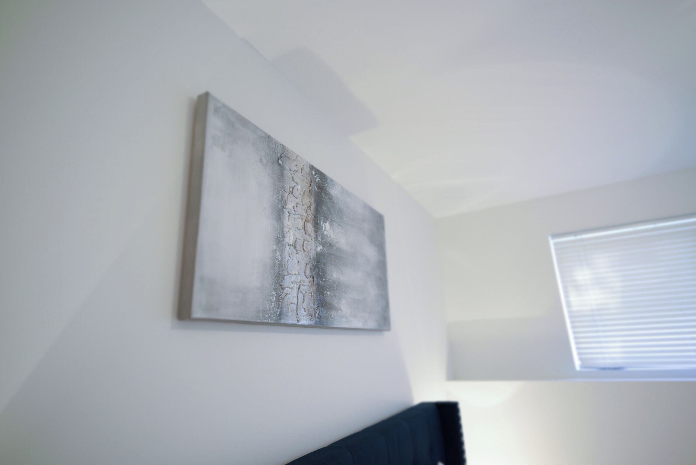 A room with a large abstract painting on the wall, a window with blinds, and a dark-colored headboard.