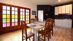 Un coin repas avec une table dressée pour six personnes, des chaises en bois, une cuisine avec des meubles blancs et une porte ornée de panneaux de vitrail colorés.