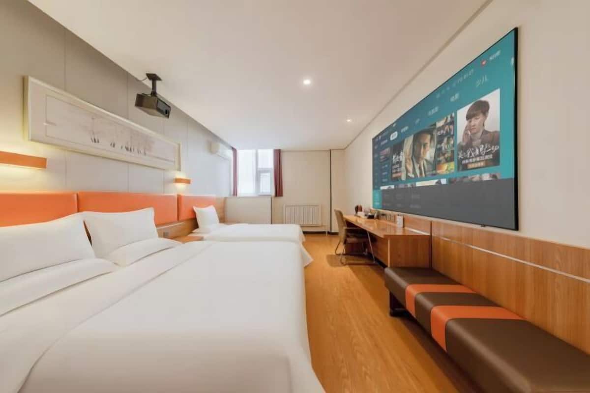 A hotel room with two beds, a desk, a seating area, a large TV screen, and a projector mounted on the wall.