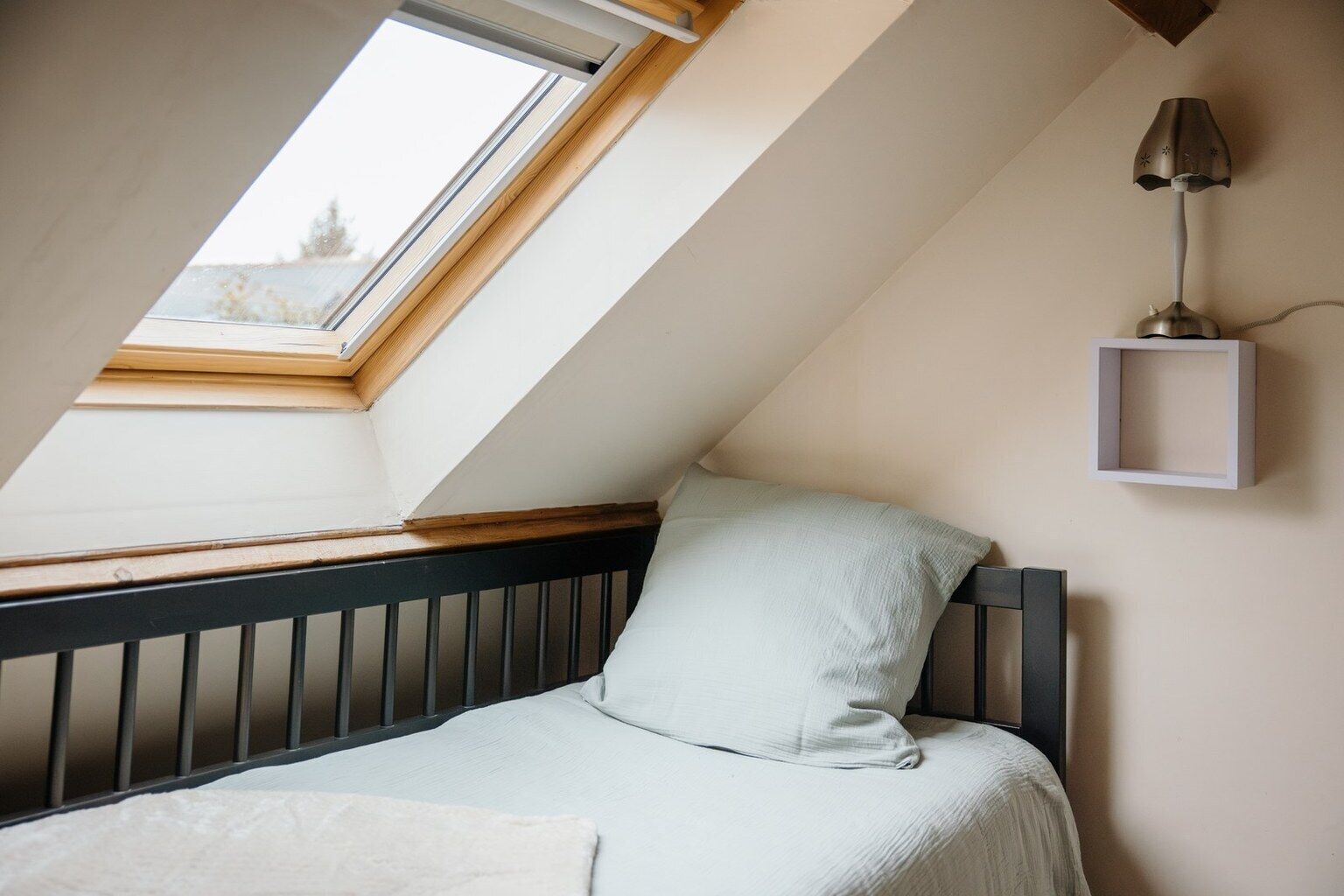 Une chambre avec un plafond en pente, un puits de lumière, un lit avec une tête de lit, une table de chevet et une lampe fixée au mur.