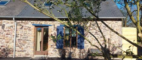 Un bâtiment en pierre avec une porte en bois et des volets bleus, un banc et un arbre au premier plan.