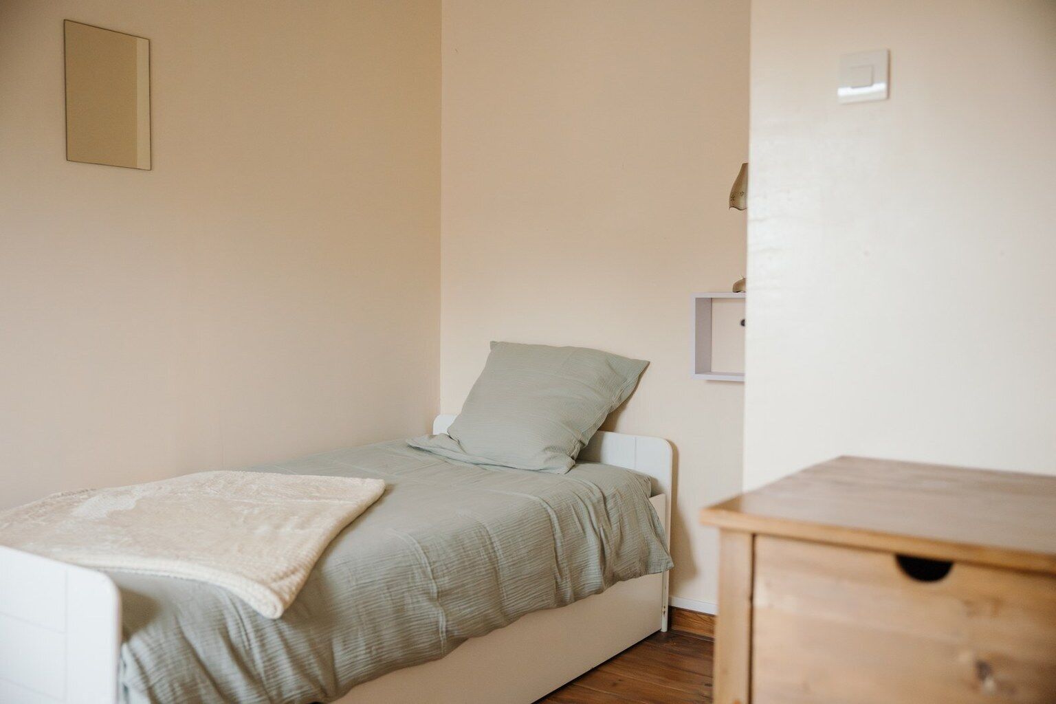 Una habitación pequeña con una cama individual, cabecera blanca, colcha verde claro y manta blanca. A la derecha de la cama, una mesita de noche de madera.