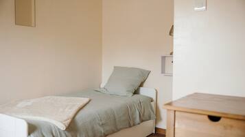 Una habitación pequeña con una cama individual, cabecera blanca, colcha verde claro y manta blanca. A la derecha de la cama, una mesita de noche de madera.