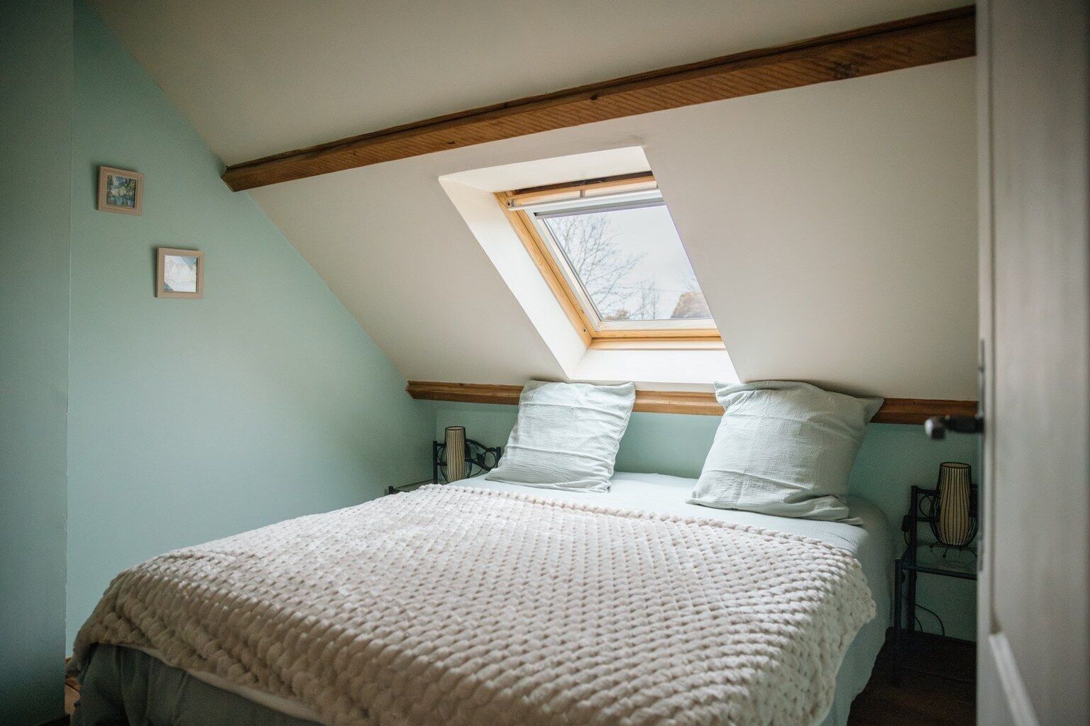 Un dormitorio con techo inclinado, un tragaluz, una cama con manta tejida, dos almohadas y una mesita de noche con lámpara.